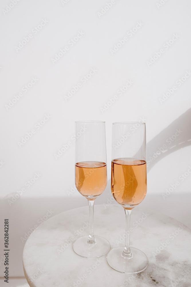 Fototapeta premium Cheers! Two glasses of rose champagne in sunlight shadows on marble tablet against white wall. Happy Birthday, anniversary party holiday celebrating decoration festive concept