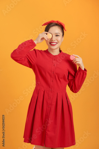 Cute Asian woman in red dress holding macaron over orange background. © makistock