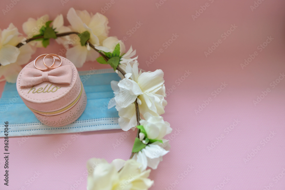 Wedding and coronavirus concept: two wedding rings, small pink box, blue medical mask (protective mask) and white flower wreath on a pink background Wedding and coronavirus concept: two wedding rings, small pink box, blue medical mask (protective mask) and white flower wreath on a pink background