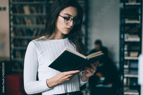 Caucasian woman in trendy eyewear for vision correction reading textbook learning for exams in university library, female student concentrated on interesting bestseller spending time on hobby
