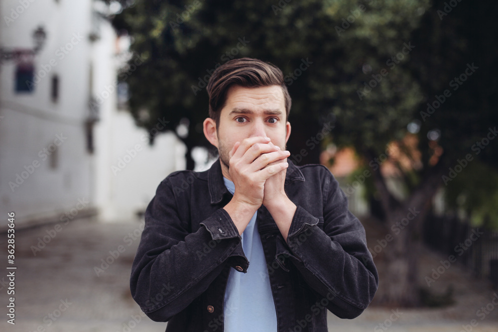 Young handsome man looks shocked, closes his mouth with his hands. A man outdoors looks scared and stressed Young handsome man looks shocked, closes his mouth with his hands. A man outdoors looks scared and stressed