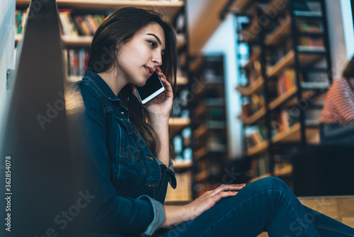 Young brunette woman in casual wear talking on cellular on free time in library with wireless connection, pretty female student calling via cellular to colleague spending time in university campus
