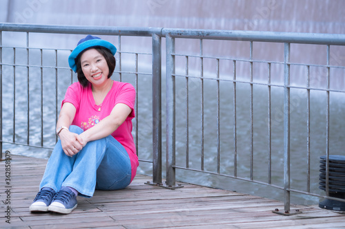 Asian happy female tourist in casual style relaxing on wooden bridge with smiling for the camera in front of waterfall decoration on building's wall in public park area