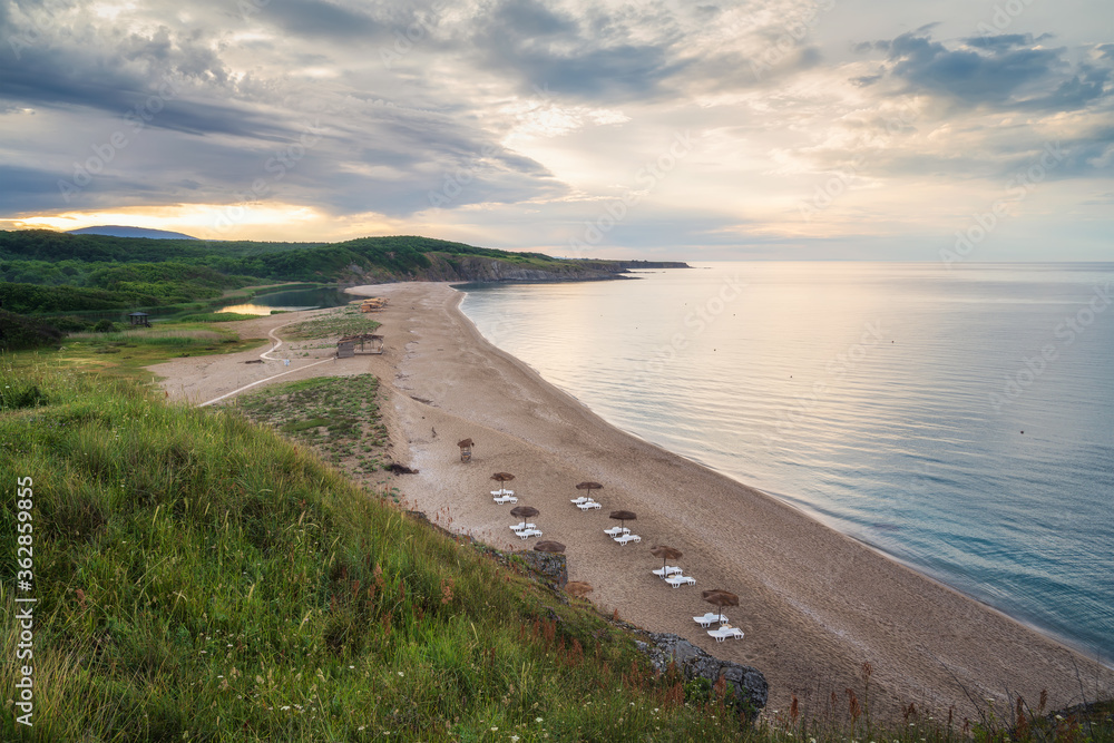 Straw sunshades and sunbeds on the empty sandy beach with sea in the background before sunset, high angle view Straw sunshades and sunbeds on the empty sandy beach with sea in the background before sunset, high angle view