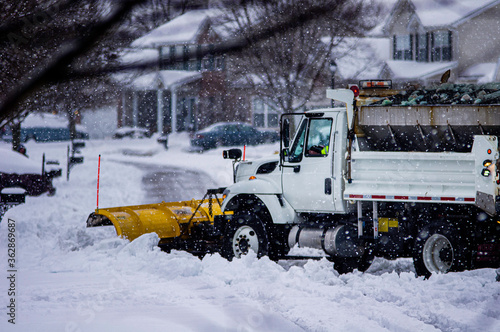 View of white and yellow city snow plow truck driving away while removing fresh deep snowfall in winter in suburban area to make roads safe for drivers