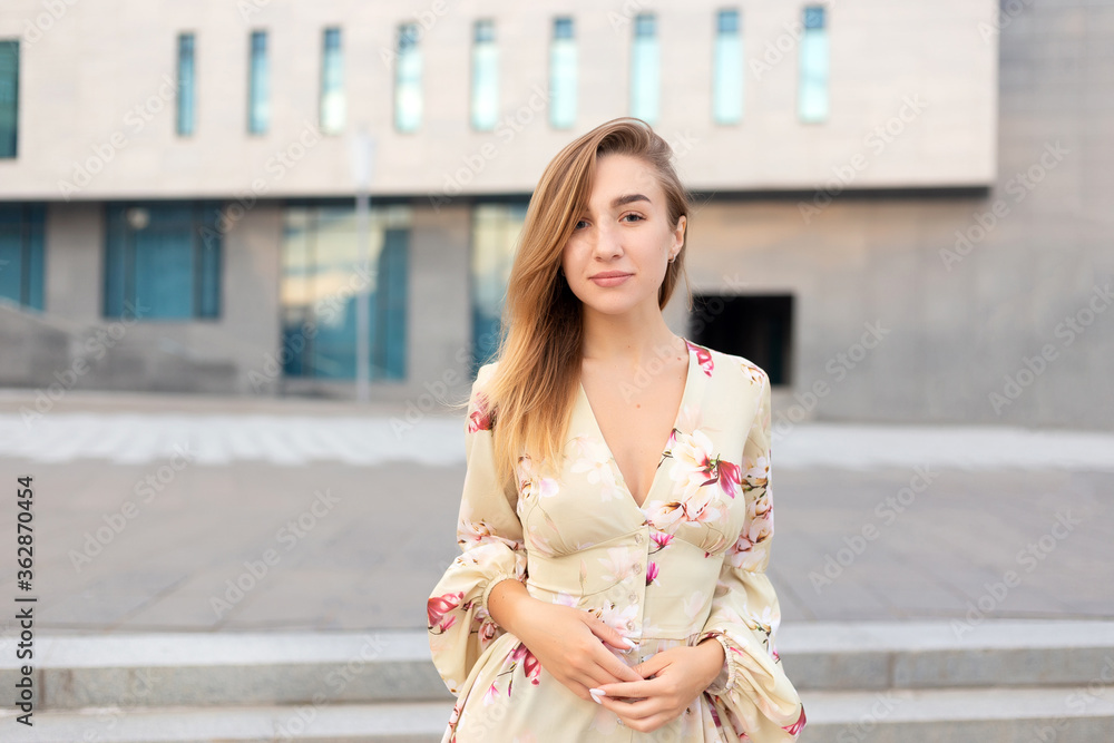Fototapeta premium Portrait of a beautiful young girl on a background of a blue business center dressed in a summer beige dress