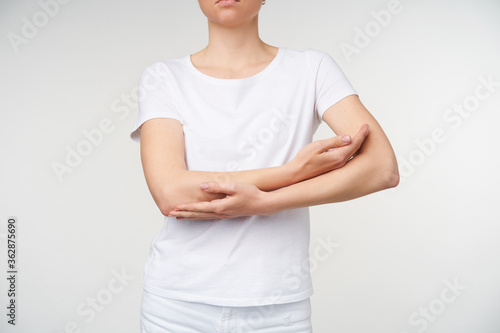 Indoor shot of young female folding together hands on her chest while imitating lulling child, standing over white background in white basic t-shirt