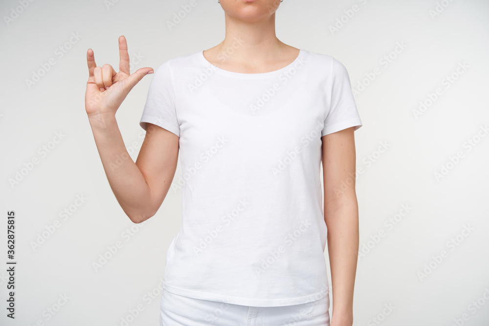 Studio photo of young woman keeping her hand raised and forming gesture while using sign language, standing over white background in casual wear Studio photo of young woman keeping her hand raised and forming gesture while using sign language, standing over white background in casual wear