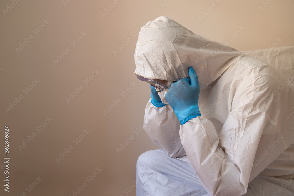 Fototapeta premium Doctor with anti-virus protective suit sitting on the bed with his hands covered in disposable gloves under his chin