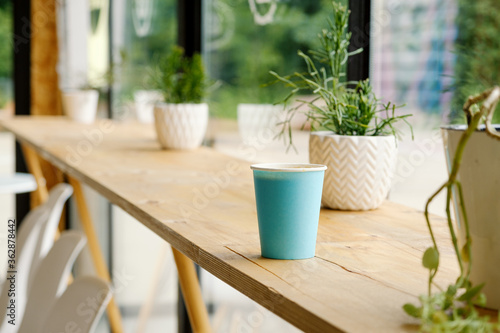 A blue paper cup with hot fresh coffee is standing on a wooden table in a cafe. The interior of the cafe with a Ripsalis cactus (twig) and other green plants. The view from the coffee shop. photo