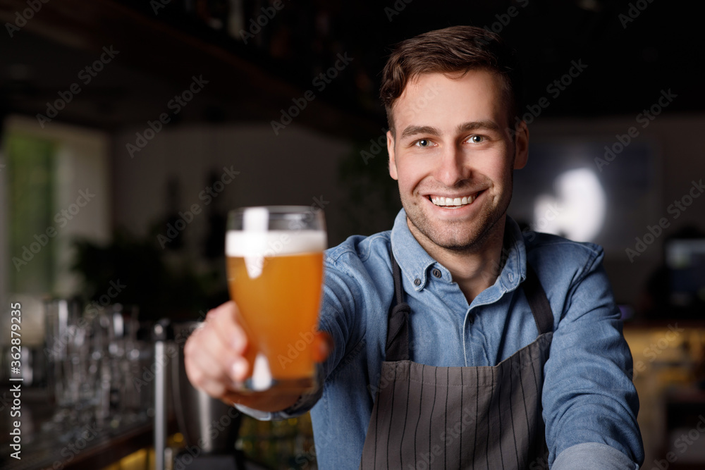 Work in pub. Smiling handsome barman in apron holds out glass of beer Work in pub. Smiling handsome barman in apron holds out glass of beer
