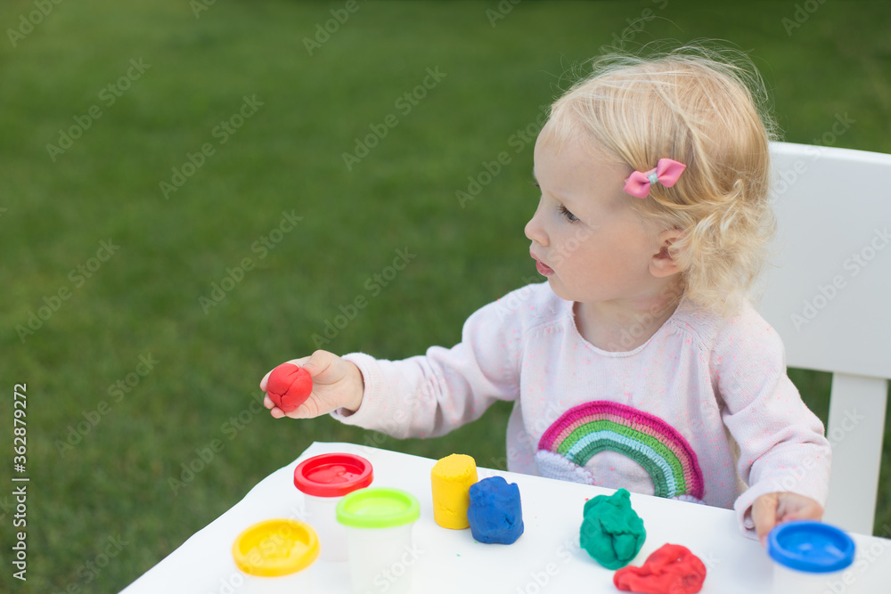 Blond toddler girl playing modeling clay in the garden in sunny summer day. She is sitting on white chair. Clay is bright and laying on white table. Blond toddler girl playing modeling clay in the garden in sunny summer day. She is sitting on white chair. Clay is bright and laying on white table.