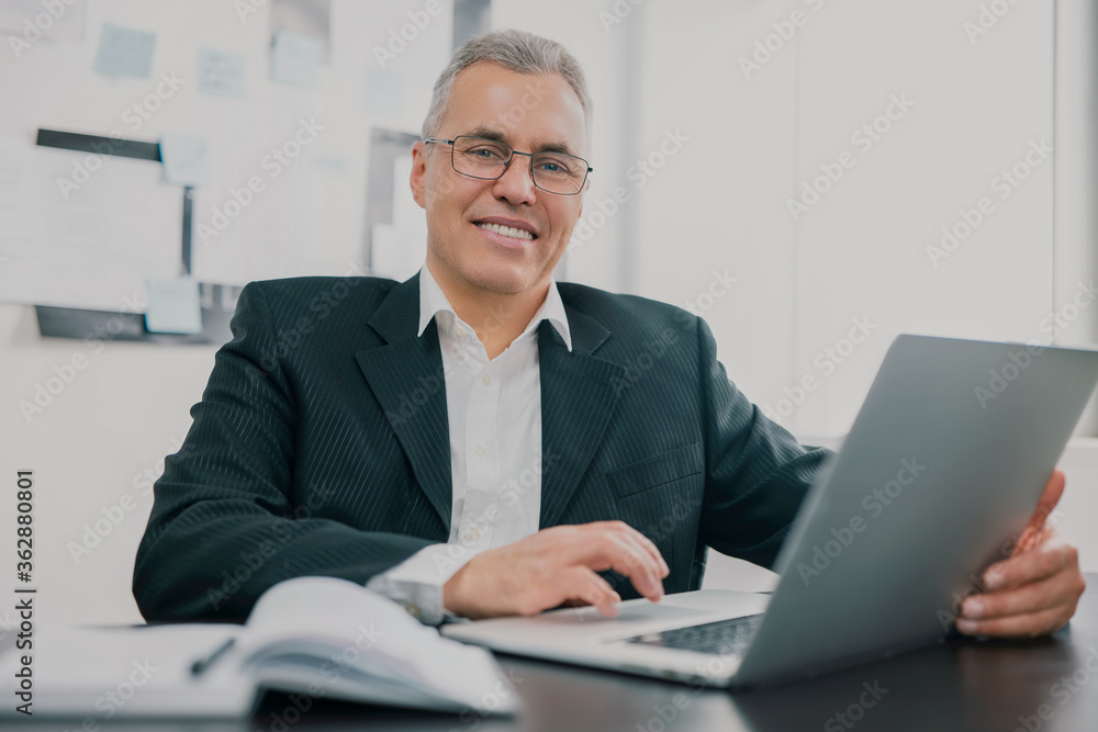 handsome gray-haired businessman in elegant suit sits in office looking excited while working in lap top on new business project , money making motivation concept handsome gray-haired businessman in elegant suit sits in office looking excited while working in lap top on new business project , money making motivation concept