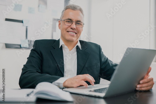 handsome gray-haired businessman in elegant suit sits in office looking excited while working in lap top on new business project , money making motivation concept