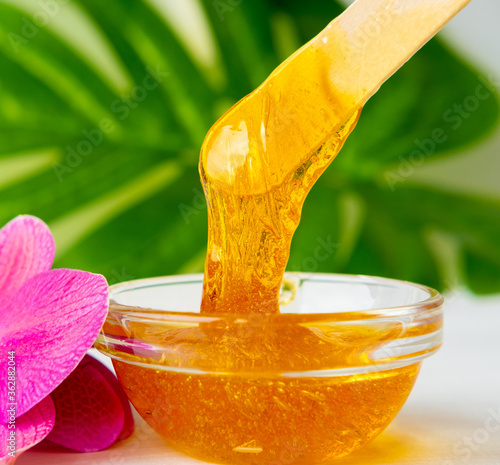 depilation and beauty concept - sugar paste or wax honey for hair removing flows down from wooden waxing spatula sticks on flower background photo