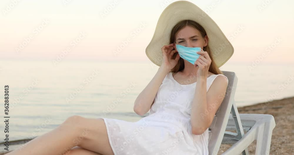 attractive young woman in a summer hat, puts a protective medical mask on her face and rests on a chaise longue on the beach. concept of summer vacation, coronavirus, quarantine attractive young woman in a summer hat, puts a protective medical mask on her face and rests on a chaise longue on the beach. concept of summer vacation, coronavirus, quarantine