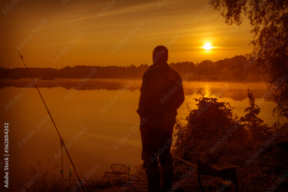 Morning on the lake dawn. A fisherman is standing on the shore next to fishing rods.