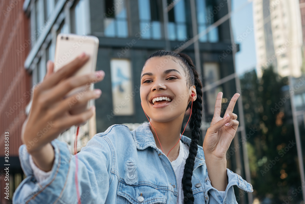 Beautiful African American woman with stylish hairstyle taking selfie, showing victory sign, standing outdoors. Young emotional blogger influencer using mobile phone, streaming video on urban street 