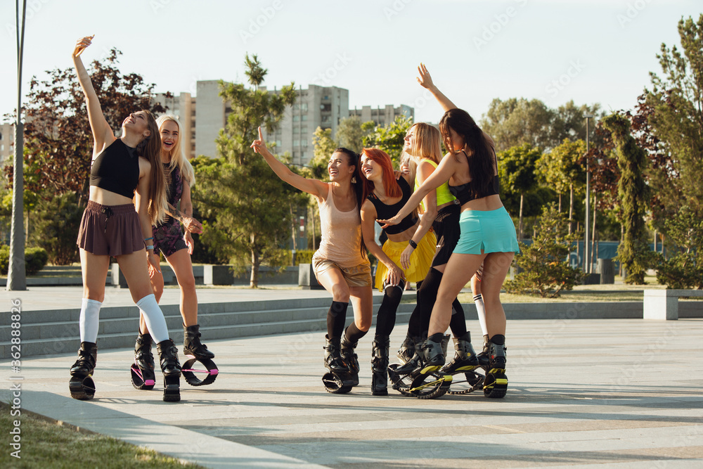 Fototapeta premium Beautiful women in sportswear jumping in a kangoo jumps shoes at the street on summer's sunny day. Jumping high, active movement, action, fitness and wellness. Fit female models during training.