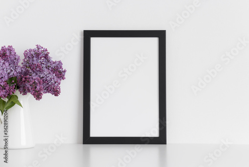 Black frame mockup with a bouquet of lilac in a vase on a white table.Portrait orientation.