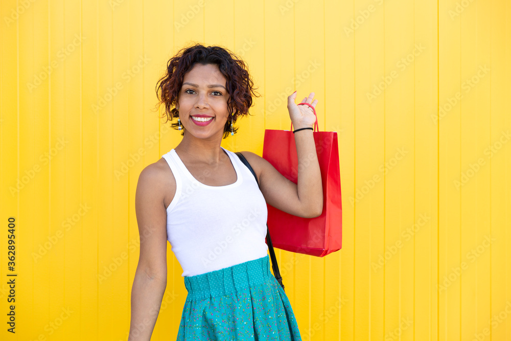 Young woman with brown skin smiling as she shows a shopping bag on a yellow wall. Space for text. Shopping, gifts and sales concept. Young woman with brown skin smiling as she shows a shopping bag on a yellow wall. Space for text. Shopping, gifts and sales concept.
