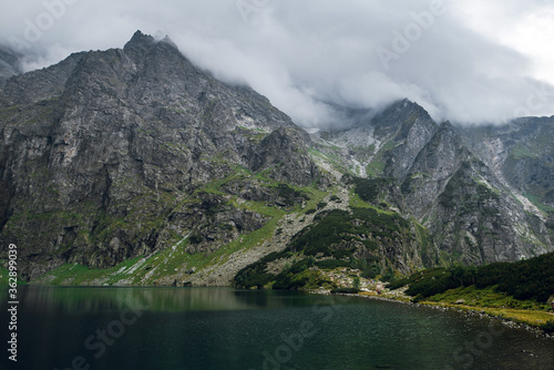 Beautiful view of foggy mountains cover by dark clouds and green forest with a reflection in a lake. Morskie Oko. Marine Eye. High Tatras, Zakopane