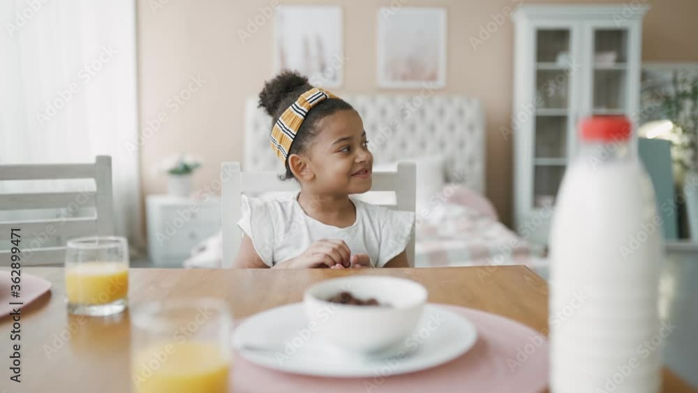 Charming little african african american girl with yellow bandage in her hair happily eats corn flakes with milk. Children's breakfast in kitchen at home before school. Good mood from morning on. Charming little african african american girl with yellow bandage in her hair happily eats corn flakes with milk. Children's breakfast in kitchen at home before school. Good mood from morning on.