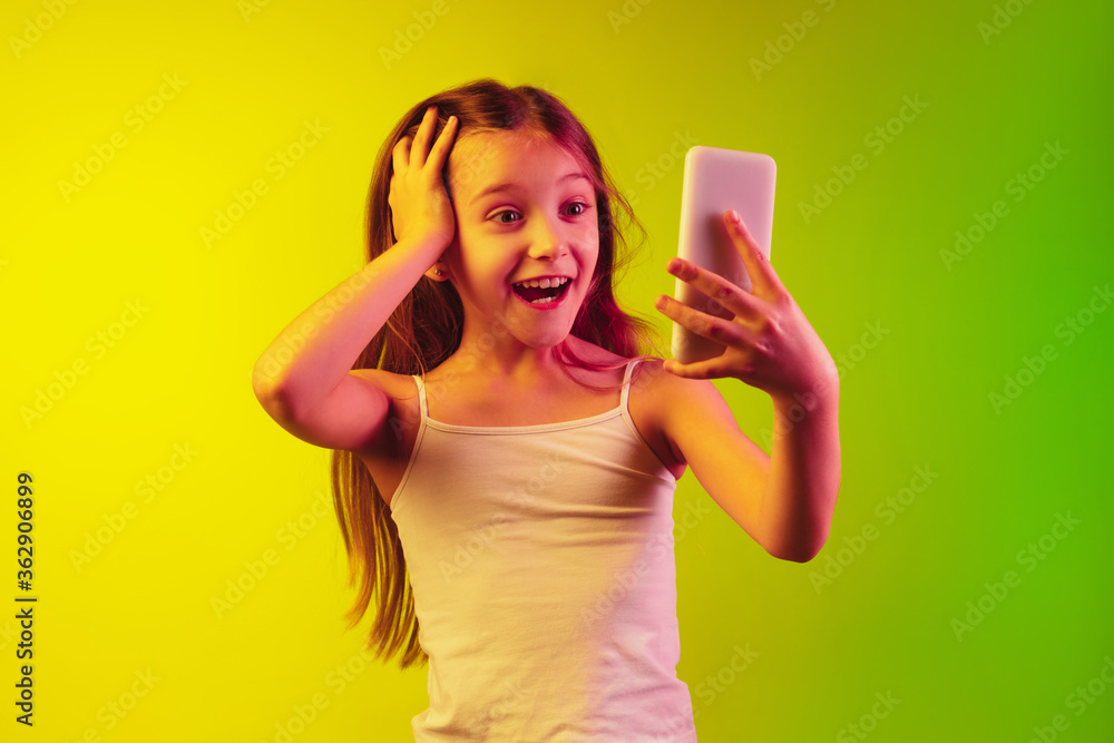 Holding phone, shocked and astonished. Little girl's portrait on gradient background in neon light. Concept of human emotions, facial expression, modern gadgets and tech, sales, ad. Copyspace. Holding phone, shocked and astonished. Little girl's portrait on gradient background in neon light. Concept of human emotions, facial expression, modern gadgets and tech, sales, ad. Copyspace.
