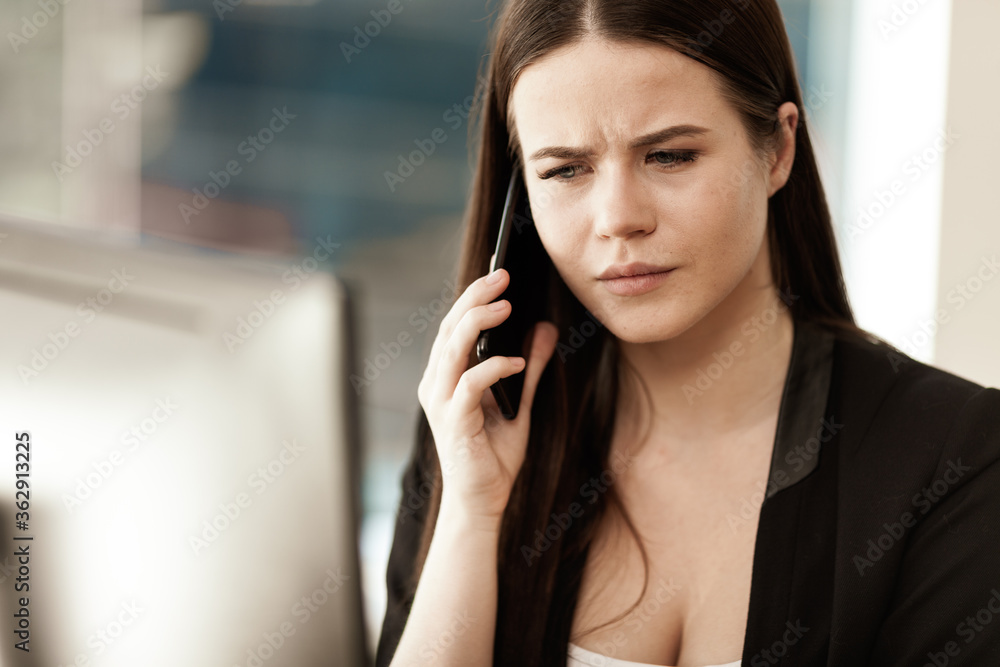 Business crisis concept. Young businesswoman sitting at the office table busy talking on a cell phone resolving a very serious work problem. Woman in suit indoors on glass window background Business crisis concept. Young businesswoman sitting at the office table busy talking on a cell phone resolving a very serious work problem. Woman in suit indoors on glass window background