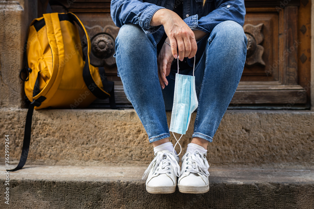 New normal at city street. Tourist woman sitting at staircase and holding face mask in hand. Travel concept during coronavirus pandemic New normal at city street. Tourist woman sitting at staircase and holding face mask in hand. Travel concept during coronavirus pandemic