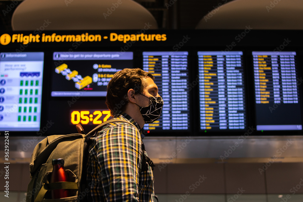Photo of a guy with a backpack looking at the Departure screen at the airport ready to catch a flight and wearing a reusable face mask.