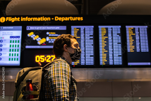 Obraz na płótnie Photo of a guy with a backpack looking at the Departure screen at the airport ready to catch a flight and wearing a reusable face mask. w salonie