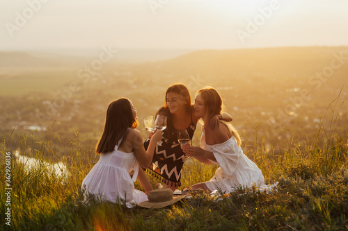 Three beautiful girls on vacation having fun, while drinking wine and eating fruits outdoors on picnic. Copy space.