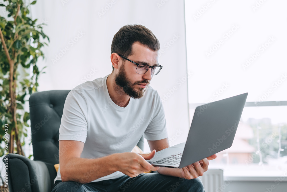 Young caucasian bearded man, freelance worker, sitting on a chair and using laptop computer. Work at home.