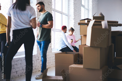 Group of young hipsters male and female colleagues having friendly conversation during break while unpacking cardboard boxes with goods from delivery service move to new loft interior coworking office