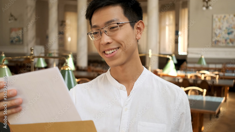 Portrait of asian student happily opening envelope with exam results in university library. Young attractive guy opening envelope in college Portrait of asian student happily opening envelope with exam results in university library. Young attractive guy opening envelope in college
