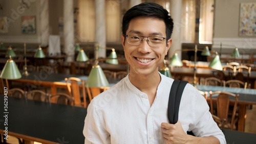 Portrait of cheerful asian student with backpack happily looking in camera in library. Happy expression