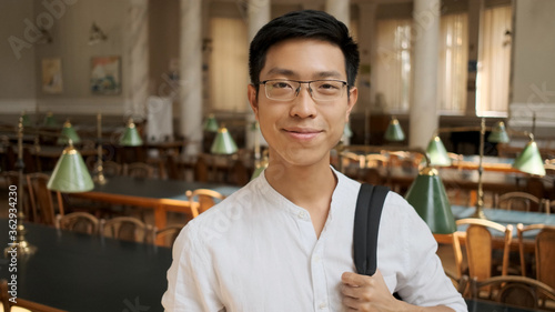 Portrait of smiling asian student with backpack happily looking in camera in college library. Young guy standing in university photo