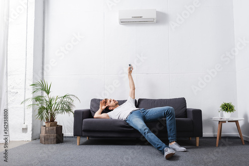 exhausted man holding remote controller and suffering from heat with broken air conditioner at home photo