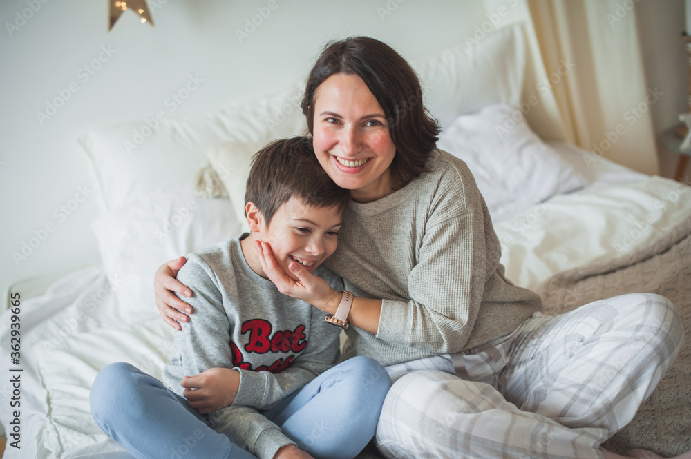 Mom and son are sitting on the bed in the bright bedroom and are smiling happily. Woman hugs teenager boy tightly. The concept of a Christmas morning, scandy style, home comfort, mom and son. Mom and son are sitting on the bed in the bright bedroom and are smiling happily. Woman hugs teenager boy tightly. The concept of a Christmas morning, scandy style, home comfort, mom and son.