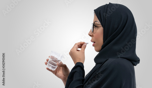 Unhealthy Arabic woman with pained facial expression feels unwell, suffering from migraine headache, takes pill analgesic medication to relieve pain, stressed lady holds tablet and glass of water.