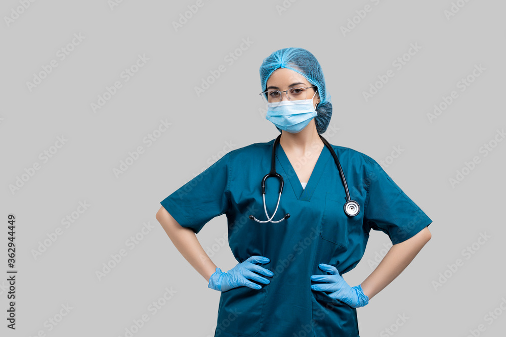 Medical doctor woman in blue uniform with stethoscope wearing surgical mask and hair cover with hands on hips standing on grey background. Medical doctor woman in blue uniform with stethoscope wearing surgical mask and hair cover with hands on hips standing on grey background.