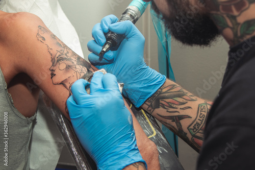 A tattoo artist paints a client's tattoo with ink. Tattoo artist holding a tattoo machine in Blue Sterile Gloves.