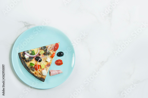 Slice of meat pizza with vegetables: , cucumber, Feta cheese, Spanish onion on black dough. cherry tomato, olive and slice of ham on turquoise serving plate, white marble background. 