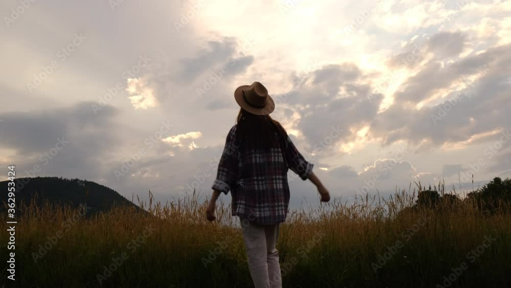 Silhouette hipster young woman in hat walks across meadow, raises hands into air with overlook amazing of mountain and sunset, beautiful and inspiring camp location for outdoor vibes living lifestyle Silhouette hipster young woman in hat walks across meadow, raises hands into air with overlook amazing of mountain and sunset, beautiful and inspiring camp location for outdoor vibes living lifestyle