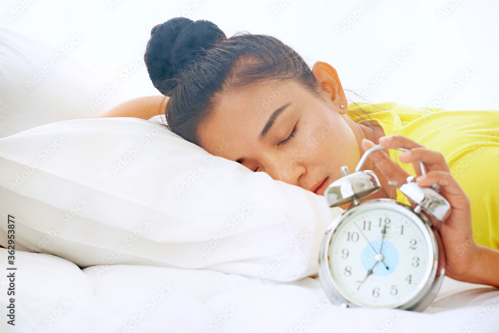 A sleepy woman lies in bed while picking up an alarm clock in the morning. - obrazy, fototapety, plakaty 