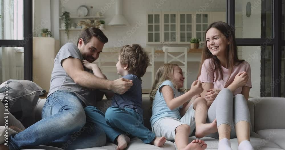 Happy joyful family mum dad with cute little kids son daughter tickling laughing sit on sofa together, young parents and small children having fun playing at home enjoy funny lifestyle game on couch Happy joyful family mum dad with cute little kids son daughter tickling laughing sit on sofa together, young parents and small children having fun playing at home enjoy funny lifestyle game on couch