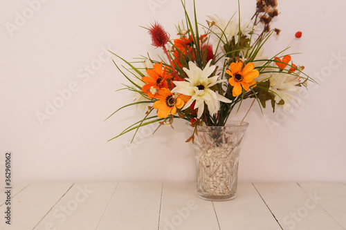 A glass vase with colorful flowers in it. Placed on top of a white wood table next to a white wall. Taken inside with studio bright lights. photo