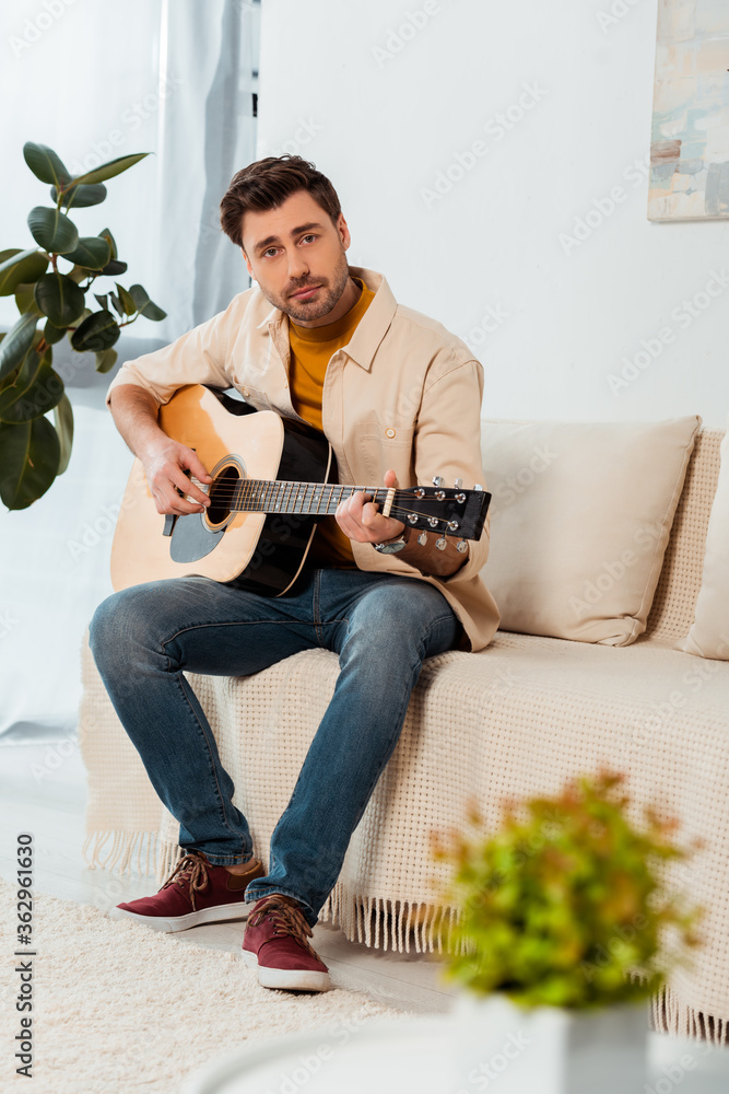 Selective focus of man looking at camera while playing acoustic guitar at home