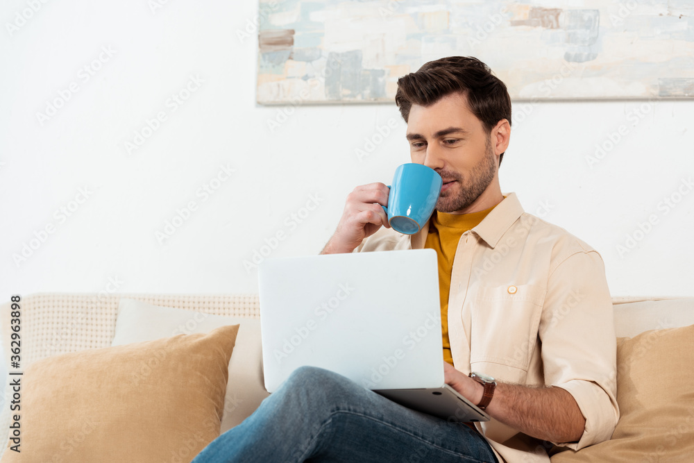 Handsome freelancer drinking coffee while using laptop in living room Handsome freelancer drinking coffee while using laptop in living room